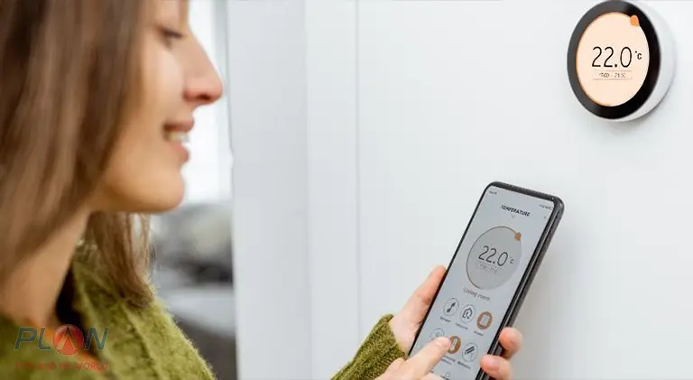 خانمی با لباس سبز یک گوشی در دست دارد و با آن دمای خانه را از طریق ترموستات هوشمند کنترل میکند.
